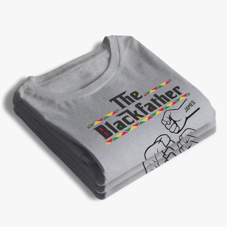May include: A folded gray t-shirt with the text "The Blackfather" in black and red, with a colorful stripe design. The shirt also features a graphic of two fists bumping and the name "JAMES".
