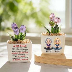 May include: Two small, white ceramic pots with glass flower sculptures. One pot has purple tulips and text in French. The other pot has pink tulips and illustrations of a mother and daughter with the names "Maman" and "Michela".