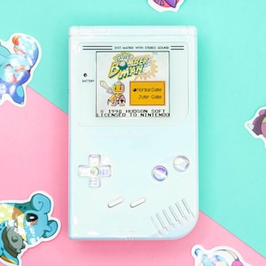 Puede incluir: Una consola de juegos portátil azul claro con el juego "Bomber Man" en la pantalla. La consola tiene una cruceta, botones y el texto "DOT MATRIX WITH STEREO SOUND". Pegatinas coloridas de personajes de dibujos animados rodean la consola.