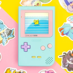 May include: A pale blue Game Boy Pocket console with a pink screen showing Pikachu surfing on waves. Various colourful Pokémon stickers surround the console on a pink and yellow background.