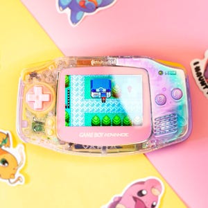 Puede incluir: Una consola portátil Game Boy Advance transparente con una pantalla rosa que muestra una escena de videojuego. La consola tiene un mando en cruz y botones rosas. Hay pegatinas de personajes de dibujos animados alrededor de la consola sobre un fondo rosa y amarillo.