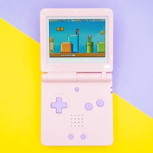 Puede incluir: Una consola de videojuegos portátil rosa, que muestra un juego de plataformas clásico en su pantalla. La consola tiene un mando direccional, botones y una pantalla. El fondo está dividido diagonalmente, con colores amarillo y morado.