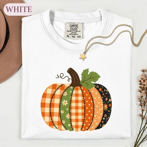 May include: A white short-sleeve t-shirt featuring a graphic of a pumpkin composed of various patterned segments including orange gingham, green floral, orange polka dot, and black floral prints, with a brown stem and green leaf.