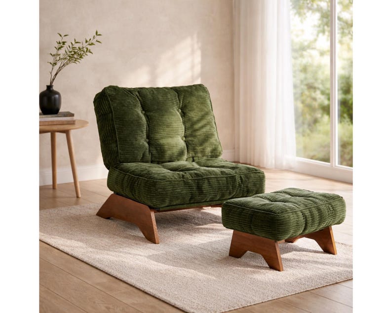 May include: A green corduroy armchair and matching ottoman with wooden legs sit on a neutral rug. The chair has a tufted back and seat. A small side table with a black vase and greenery is in the background.