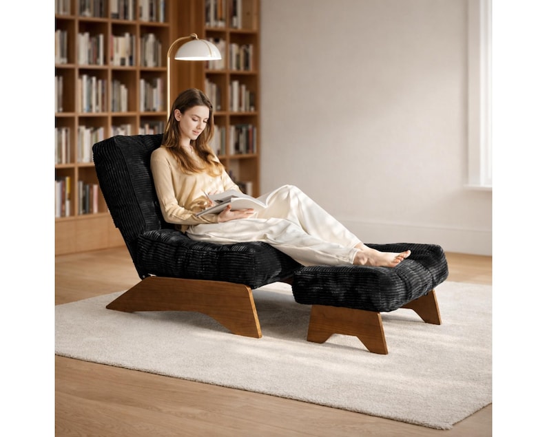 May include: A black corduroy lounge chair with a matching ottoman, both with wooden legs. A person is seated in the chair, reading a book. A white lamp with a gold arm is positioned above the chair. The chair and ottoman are on a light-colored rug.