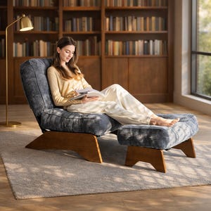 May include: A blue corduroy armchair and matching ottoman with wooden legs. The chair is angled, and the ottoman is placed in front. A person is sitting in the chair, reading a book. A gold floor lamp is to the left.