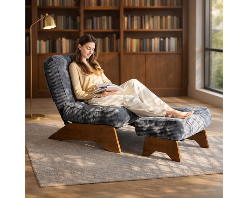 May include: A blue corduroy armchair and matching ottoman with wooden legs. The chair is angled, and the ottoman is placed in front. A person is sitting in the chair, reading a book. A gold floor lamp is to the left.