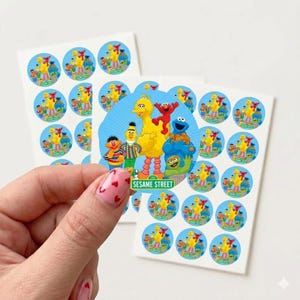 May include: A sheet of round stickers featuring Sesame Street characters like Big Bird, Elmo, and Cookie Monster. The stickers have a blue background and a white border. The image includes a hand holding a sticker.