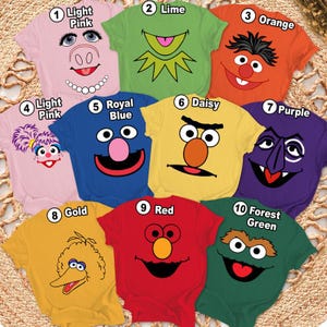 May include: A collection of ten colorful t-shirts, each featuring a different cartoon character face. The shirts are in various colors, including light pink, lime, orange, royal blue, daisy yellow, purple, gold, red, and forest green.