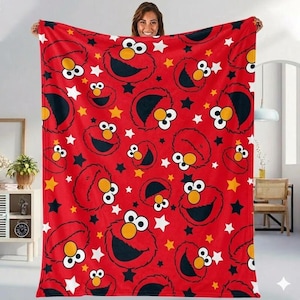Puede incluir: Manta roja con un patrón repetido de la cara de Elmo, estrellas negras y estrellas blancas. La manta está sostenida por una persona. La manta es un artículo suave y acogedor.