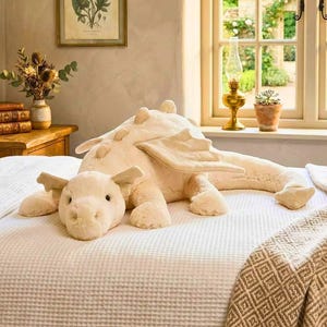 May include: A large, cream-coloured plush dragon toy with wings and a smaller dragon head, resting on a white waffle-weave bedspread. A framed botanical print and a window with a garden view are in the background.