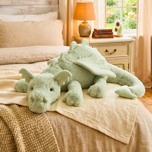 May include: A large, soft, mint green dragon plush toy with wings, lying on a bed with a beige blanket. The dragon has a friendly face and is surrounded by a cozy bedroom setting with a lamp and a window.