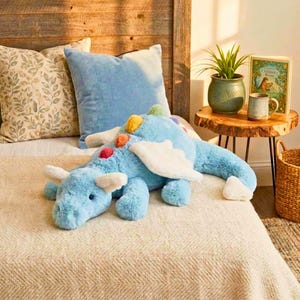 May include: A soft, light blue dragon toy with white wings and colourful accents. The stuffed animal is on a bed with pillows, a wooden headboard, and a side table with a plant and a book.