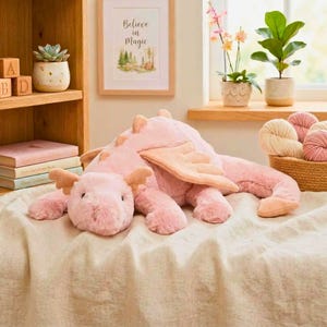 May include: A large, plush pink dragon toy with soft wings and a friendly face. The dragon is lying on a cream-colored blanket. The background includes a wooden shelf with books, a framed picture that says "Believe in Magic", and potted plants.