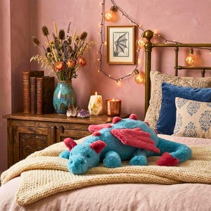 May include: A plush blue dragon toy with pink wings and accents rests on a cream-colored blanket atop a bed. The dragon is the focal point, with a cozy bedroom setting in the background, including a wooden nightstand, vase of flowers, and string lights.