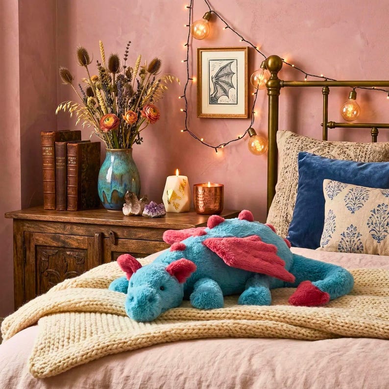 May include: A plush blue dragon toy with pink wings and accents rests on a cream-colored blanket atop a bed. The dragon is the focal point, with a cozy bedroom setting in the background, including a wooden nightstand, vase of flowers, and string lights.