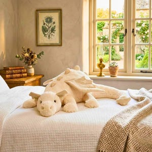 May include: A large, cream-colored plush dragon toy with wings and a smaller dragon head. The toy is lying on a white waffle-weave bedspread. A framed botanical print and a window with a garden view are in the background.