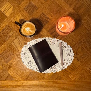 May include: A dark brown leather journal with a pen and a cup of coffee with latte art rest on a white doily. A lit candle in an orange glass sits nearby. The items are arranged on a wooden table.