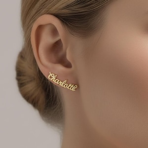 Puede incluir: Pendientes dorados con el nombre "Charlotte" escrito en cursiva. Los pendientes se llevan en la oreja de una persona con cabello rubio. Los pendientes son una joya delicada.