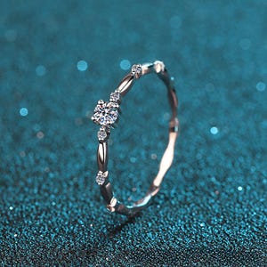 Pode incluir: Um delicado anel de prata com um diamante redondo central brilhante e pedras de acento menores. A banda apresenta um design texturizado único. O anel está sobre um fundo azul-petróleo.