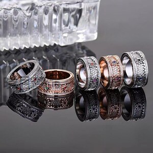 Peut inclure: Plusieurs bagues décoratives en argent, or rose et noir. Les bagues présentent des motifs complexes avec des pierres précieuses incrustées de différentes couleurs. Les bagues sont exposées sur une surface réfléchissante, avec un objet en verre transparent en arrière-plan.