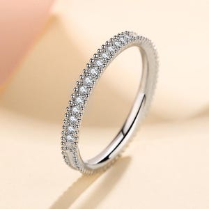 Puede incluir: Un anillo plateado con una banda de pequeñas piedras preciosas transparentes. El anillo se presenta sobre un fondo suave y neutro, resaltando su delicado diseño y brillo. La superficie pulida refleja la luz, realzando su elegancia.