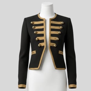 Könnte beinhalten: Schwarze Jacke mit goldenen Verzierungen und Knöpfen. Die Jacke hat einen runden Kragen und lange Ärmel. Goldene Zierleisten säumen die Kanten, Taschen und Manschetten. Goldene Knöpfe zieren die Vorderseite und die Ärmel. Die Jacke wird auf einer Schaufensterpuppe präsentiert.