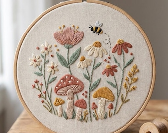 May include: A round embroidery hoop featuring a floral and mushroom design. The artwork includes various flowers, mushrooms, and a bee, all in shades of pink, red, yellow, and green. The hoop is made of wood and the fabric is a light beige color.