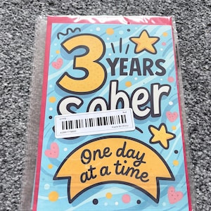 May include: A greeting card celebrating three years of sobriety. The card features the words "3 YEARS sober" in a playful font, with a banner that reads "One day at a time." The design includes stars and hearts.