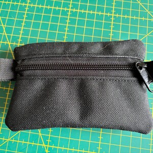Puede incluir: Pequeño estuche negro con cremallera de forma rectangular. El estuche tiene una cremallera negra y presillas de tela a cada lado. Está hecho de un material texturizado y duradero. El fondo es una esterilla de corte verde.