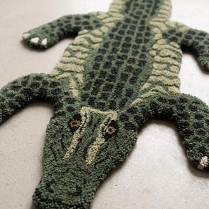 Peut inclure: Un tapis tufté en forme d'alligator, présentant une surface texturée avec une palette de couleurs vert foncé, vert olive et noir. Le motif inclut des détails d'écailles sur le dos et la tête, avec de petites griffes de couleur claire.