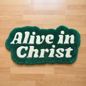 Puede incluir: Una alfombra de mechones con una forma irregular similar a una nube, con un fondo verde bosque profundo y el texto "Alive in Christ" en letras grandes, redondeadas y de color crema. La alfombra tiene una superficie de pelo grueso y texturizado.