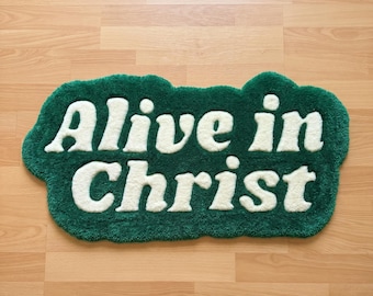 Puede incluir: Una alfombra de mechones con una forma irregular similar a una nube, con un fondo verde bosque profundo y el texto "Alive in Christ" en letras grandes, redondeadas y de color crema. La alfombra tiene una superficie de pelo grueso y texturizado.
