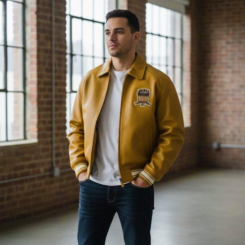 May include: Mustard yellow leather jacket with a white and navy striped cuff and collar. The jacket has a patch on the chest with a crest design. The person is wearing a white t-shirt and dark blue jeans.