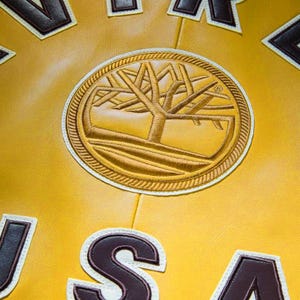 May include: Close-up of a yellow leather jacket with a large, circular patch featuring a tree design in gold and brown. The jacket has brown and white letter patches. The overall aesthetic is vintage and sporty.