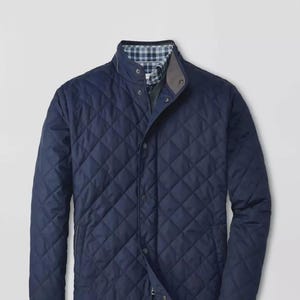 May include: A navy blue quilted jacket featuring a diamond-patterned stitch, a stand-up collar with snap button closures, and a visible blue and white plaid lining inside the neck area. The jacket includes long sleeves and a front snap placket.