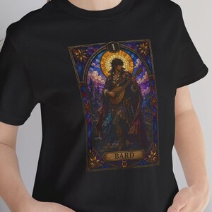 Può includere: T-shirt nera con una grafica in stile vetrata raffigurante un bardo che suona un liuto. La grafica include il numero "1" e la parola "BARD". Il design ha un tema fantasy medievale.