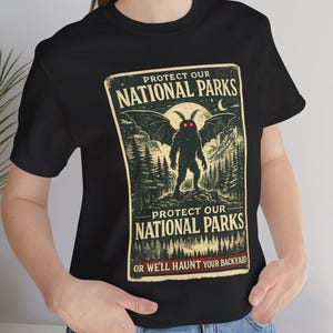 Peut inclure: T-shirt noir avec un motif vintage. Le design comprend le texte "PROTECT OUR NATIONAL PARKS" et une créature ailée aux yeux rouges, sur fond de forêt. La phrase "OR WE'LL HAUNT YOUR BACKYARD" est également présente.