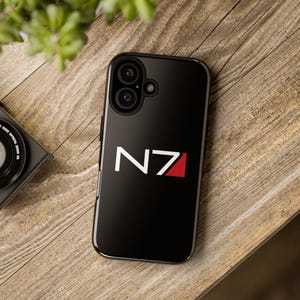 Peut inclure: Coque de smartphone noire avec les lettres blanches "N7" et une bande rouge en biais. La coque est posée sur une surface en bois, avec un appareil photo et une plante en arrière-plan.