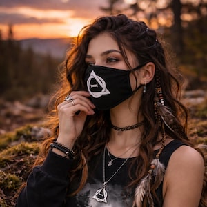 May include: Black face covering featuring a white geometric symbol. The person wearing the mask has long, wavy brown hair and is wearing a black top and necklaces. The background shows a sunset.