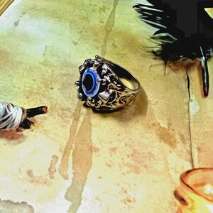 May include: A silver-toned metal ring featuring an intricate, textured band with a central, circular blue and white eye-shaped inlay. The eye design has a dark pupil surrounded by concentric blue and white rings, set within a detailed metal frame.