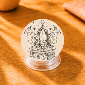 Op de afbeelding: Een crèmekleurige popsocket met een zwart-wit illustratie van een fantasie scène. Het ontwerp toont draken, een boog, boeken en kristallen. De popsocket heeft een transparante plastic basis.