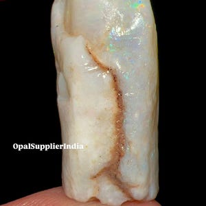 Puede incluir: Una piedra preciosa de ópalo blanco en bruto con destellos iridiscentes de color. El ópalo tiene una veta marrón que lo atraviesa. El texto "OpalSupplierIndia" es visible en la imagen.