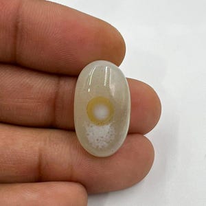 Può includere: Una gemma cabochon di forma ovale e traslucida con una superficie liscia e lucida. La pietra presenta un colore di base beige lattiginoso pallido con un distinto motivo circolare centrato composto da un nucleo bianco circondato da un anello giallo dorato.