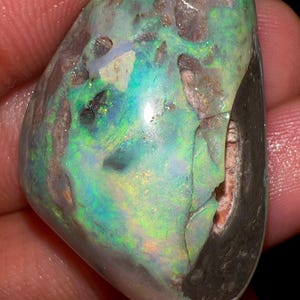 May include: A polished opal gemstone with a mix of colors, including green, blue, and hints of brown. The stone has a unique, irregular shape and a cracked surface. The opal is held in a hand.