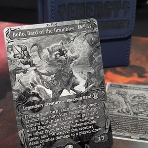 May include: A black and white Magic: The Gathering card featuring "Bello, Bard of the Brambles," a raccoon bard. The card displays a detailed illustration of the character and game text. The card is propped up in front of a blue card case.