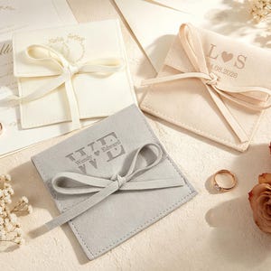 Puede incluir: Tres bolsas cuadradas de terciopelo para joyas en blanco, crema y gris, cada una con un cierre de lazo. La bolsa crema está grabada con L corazón S y 04.08.2026, mientras que la bolsa gris muestra el texto Wendy corazón Edward.