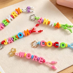 Puede incluir: Cinco llaveros personalizados con cuentas de letras de plástico de colores que forman los nombres HENRY, ANNA, JAXON, COCO y WORE. Cada llavero incluye pequeñas cuentas redondas, un cierre metálico en forma de flor o lazo y extremos anudados.