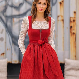 Könnte beinhalten: Ein rotes Dirndlkleid mit quadratischem Ausschnitt und eng anliegendem Oberteil. Das Kleid hat ein weißes Spitzentop und lange Ärmel. Ein Schleifendetail akzentuiert die Taille. Das Kleid hat einen Reißverschluss und einen weiten Rock.