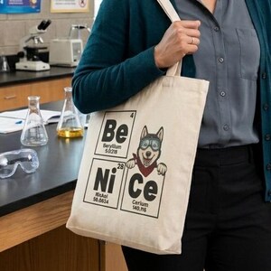 Puede incluir: Una bolsa de mano beige con las palabras "Be Ni Ce" y un husky de dibujos animados con un pañuelo rojo. La bolsa la lleva una persona en un aula de ciencias con tablas periódicas y equipos de laboratorio en el fondo.